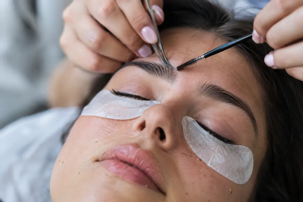 Stylistka stylizuje brwi klientki, pod oczami hydrożelowe płatki